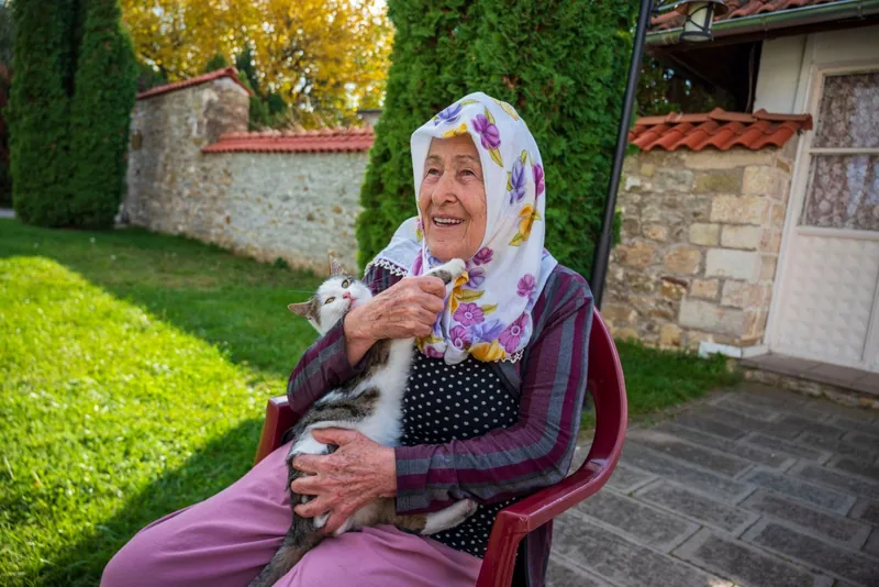 A smiling older woman holding a cat in a sunny garden